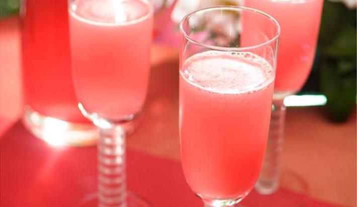 Fotografia em tons de rosa em uma bancada de madeira, um pano vermelho, taças de vidro de champagne com as bebidas feitas com Picolé de Limão com espumante.