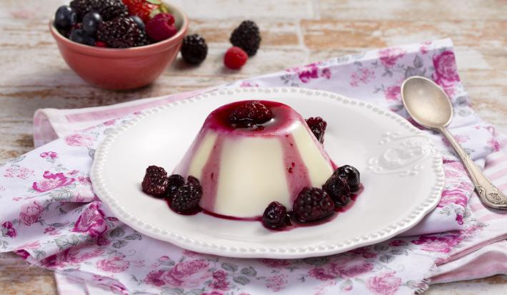 fotografia em tons de branco e roxo, contém um pano branco com rosas desenhadas, acima um prato redondo branco com um mini Flan com calda de frutas vermelhas e frutas vermelhas em volta.