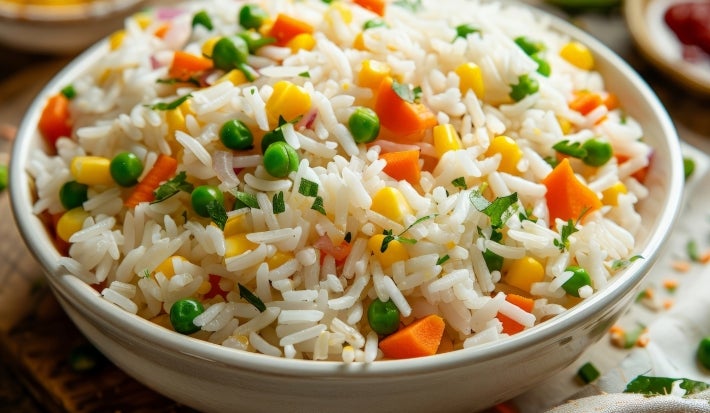 Em cima de uma mesa, tigela com porção de arroz branco soltinho com milho, cenouras e milho-verde, decorado com fatias de bacon e folha de salsa.
