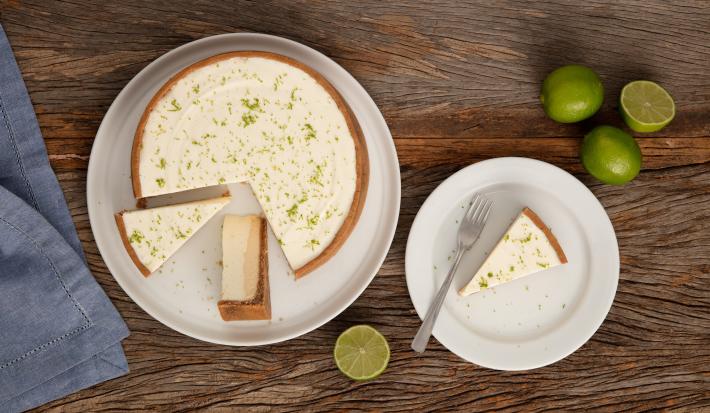 Fotografia em tons de branco e marrom de uma bancada de madeira vista de cima, com um prato branco com uma torta de limão fatiada, ao lado direito prato com uma porção de torta com um garfo e limões, e no esquerdo um guardanapo azul.