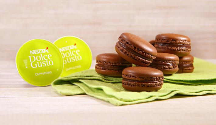 Fotografia em tons de verde em uma bancada de madeira clara, um paninho verde com vários macarons de chocolate e café em cima dele. Ao lado, cápsulas de Cappuccino Dolce Gusto.
