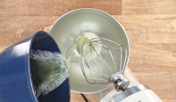 Panela despejando mistura cozida de claras e açúcar dentro do bowl de inox de uma batedeira.