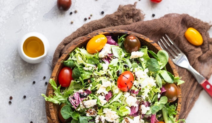 Fotografia de uma salada em uma tigela de madeira, repleta de folhas verdes, tomates-cereja coloridos e queijo branco esfarelado, com um toque de molho balsâmico. Um pequeno recipiente de azeite e tomates inteiros decoram a cena.