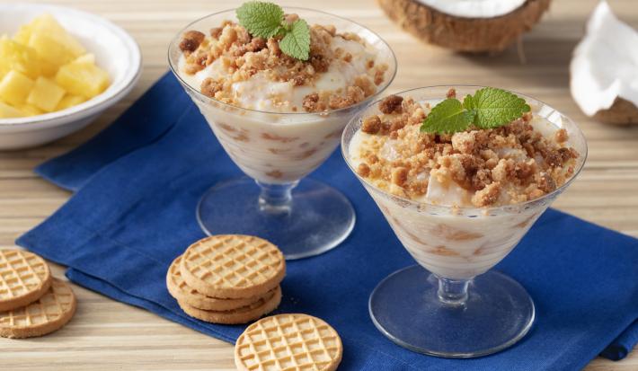 Fotografia em tons de marrom e azul de uma bancada de madeira com um paninho azul, sobre ele duas taças com o creme e biscoitos. Ao fundo um coco e um recipiente redondo com abacaxi.