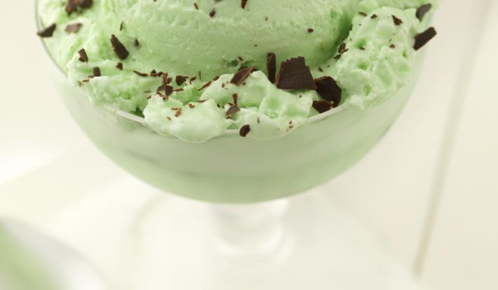 Fotografia de uma bola de sorvete de menta com chocolate em uma taça, que está sobre uma mesa branca. E, ao lado da taça, uma colher de sobremesa.