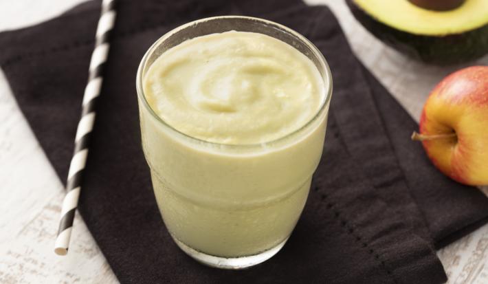 Fotografia em tons de branco e preto de uma bancada de madeira branca com um paninho preto, sobre ele um copo de vidro com o creme de abacate e um canudo. Ao fundo meio mamão e uma maçã.