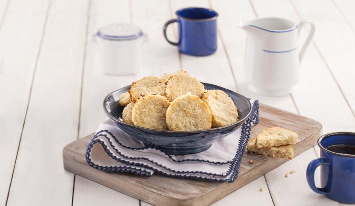 Fotografia em tons de azul em uma bancada de madeira branca, uma tábua de madeira, um paninho branco com detalhes azuis, um recipiente azul redondo fundo com os biscoitos de coco com amêndoas dentro. Ao lado, uma xícara azul.
