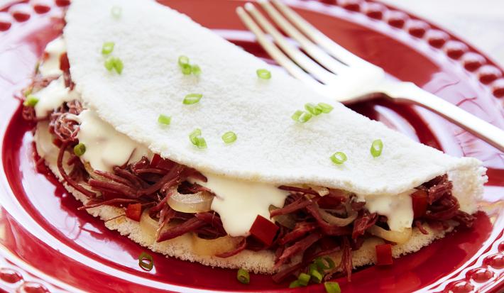 Fotografia em tons de branco e vermelho, com prato vermelho ao centro contendo uma tapioca com recheio de carne-seca, queijo de coalho e salpicada de cebolinha, acompanhada de um garfo, sobre guardanapo multicolorido.