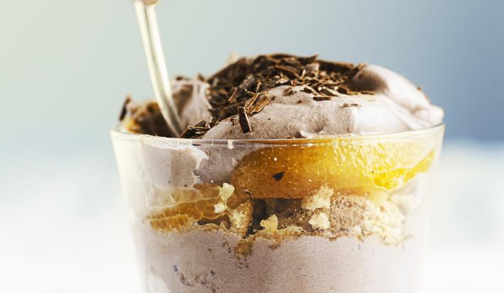 Fotografia de um copinho de vidro, dentro tem pavê com biscoitos champanhe, laranja em gomos e creme de chocolate meio amargo. Por cima do pavê tem raspas de chocolate. O copo está sobre uma toalha de mesa branca.