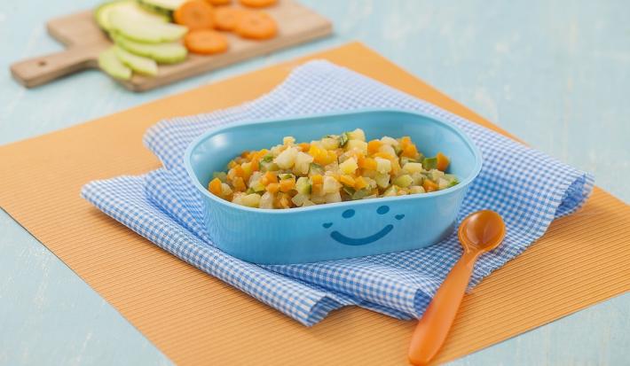 Fotografia em tons de laranja em uma bancada de madeira de cor azul. Ao centro, um pano laranja e um pano quadriculado azul, em cima, há um recipiente contendo os legumes e ao lado, uma colher laranja. Ao fundo, uma tábua contendo alguns legumes.