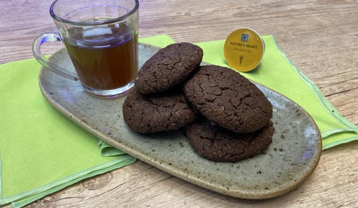 Fotografia em tons de verde em uma bancada de madeira, um recipiente oval marrom ao centro com 4 cookies de chocolate com amêndoas e ao lado uma xícara de vidro com o chá mate com acerola. Ao lado, uma cápsula de Dolce Gusto.