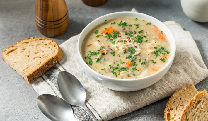 Imagem de um bowl branco com a receita de canja de galinha decorada com salsinha. Ao lado, pedaços de pão e duas colheres.