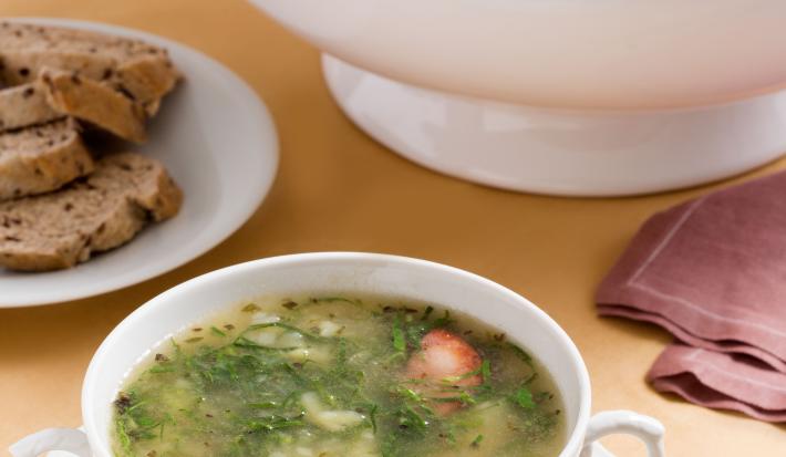 Foto de um recipiente redondo, como uma xícara, só que maior, com caldo verde dentro, em cima de um prato branco