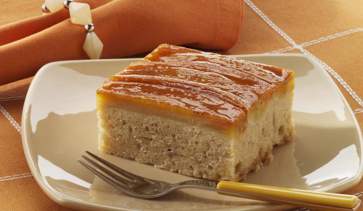 Foto de um prato branco em cima de uma toalha laranja com branco e com um pedaço de bolo de banana dentro com um garfo do lado