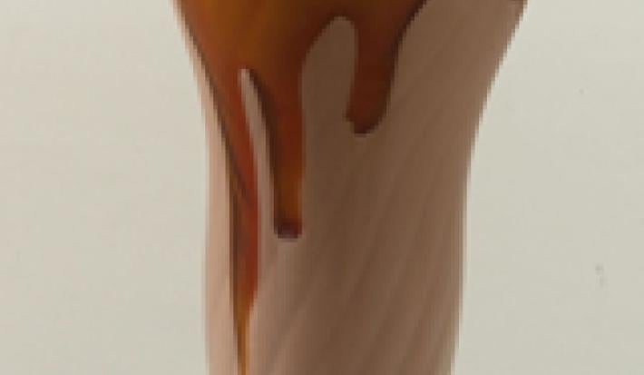 Fotografia em tons de marrom em uma bancada cinza com uma taça de vidro alta e o milkshake de chocolate dentro dela.