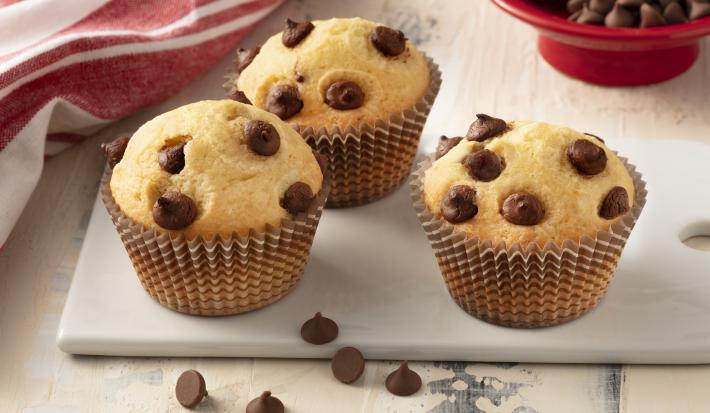 Fotografia em tons de branco e vermelho de uma bancada branca com uma tábua banca, sobre ela três muffins. Ao fundo um paninho vermelho e um recipiente redondo vermelho com gotas de chocolate.