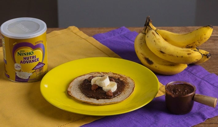 Fotografia de uma mesa de madeira com dois panos, roxo e amarelo, um por cima do outro. Apoiado neles tem um prato amarelo com uma panqueca de banana e creme de avelã. Ao redor tem uma lata de NINHO Nutri Advance, um cacho de bananas e o creme.
