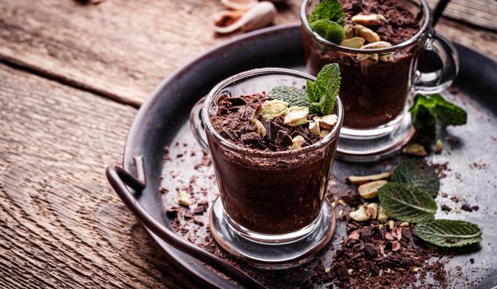 Fotografia de dois recipientes pequenos de café com um gelado de chocolate e nozes, com folhas de hortelã e mais nozes por cima. Os recipientes estão sobre uma decoração preta parecida com uma panela de ferro, com folhas de hortelã, nozes e chocolate.