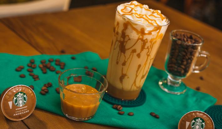 Fotografia em tons de verde de uma mesa de madeira com um paninho verde e ao centro um copo decorado com doce de leite, com uma bebida de leite e decorado com chantilly. Ao redor cápsula e grãos de café. Fotografia em tons de verde de uma mesa de madeira com um paninho verde e ao centro um copo decorado com doce de leite, com uma bebida de leite e decorado com chantilly. Ao redor cápsula e grãos de café.