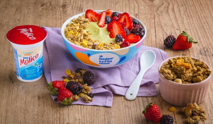Fotografia em tons de lilás em uma bancada de madeira clara, um bowl colorido com o iogurte de abacate com frutas vermelhas e granola decorando. Ao lado, um potinho com granola e frutas espalhadas na bancada.