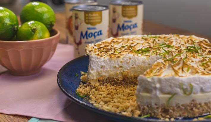 Foto de uma bancada, sobre ela há uma prato azul com a receita da Torta de Limão Fácil sem uma fatia, um pote rosa com limões verdes e algumas latas de Leite Moça ao fundo