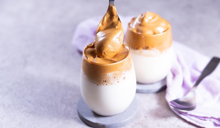 Fotografia de dois copos de vidro com cappuccino cremoso, com espuma de chocolate por cima, também cremoso. Cada copo está apoiado em um apoio cinza, o qual está sobre uma mesa de cimento queimado.