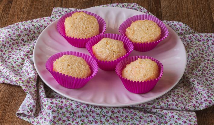 Imagem da receita de Queijadinha da Miriam Braga, servida em forminhas rosas de cupcakes, sobre um prato rosa, em um bancada de madeira decorada com um tecido florido