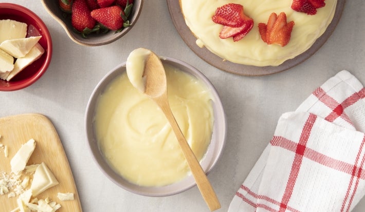 Imagem vista de cima de um bowl com a ganache de chocolate branco e uma colher de madeira. Ao lado, um bolo coberto com a ganache, morangos e pedaços de chocolate branco. Imagem vista de cima de um bowl com a ganache de chocolate branco e uma colher de madeira. Ao lado, um bolo coberto com a ganache, morangos e pedaços de chocolate branco.