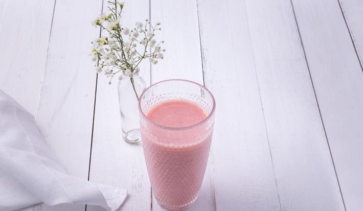 Fotografia em tons de rosa em uma bancada de madeira clara com um copo de vidro ao centro e a vitamina de hortelã com goiaba dentro dele. Ao fundo, um vasinho de vidro com flores brancas pequenas.
