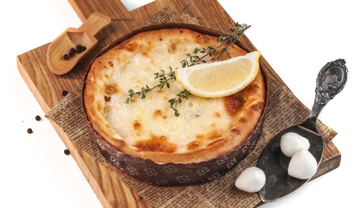 Fotografia de um bolo de mandioquinha levemente gratinado, com alecrim e um pedaço de limão siciliano por cima. O bolo está apoiado sobre uma folha de papel manteiga desenhado, que está em cima de uma tábua de madeira. O fundo da foto é branco.
