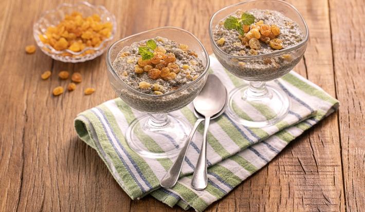 Foto da receita de Pudim de Chia com Uva Passa. Observa-se duas taças de vidro transparentes com o pudim e as uvas, sobre um guardanapo listrado em tons de verde claro. Também há duas colheres sobre o guardanapo e, atrás, há um pote com uvas passas.