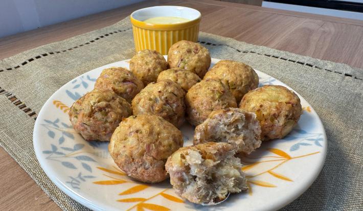 Fotografia em tons claros de um prato raso médio branco sobre um paninho em tom pastel sobre uma mesa de madeira. Dentro do prato tem várias unidades do bolinho de atum com batata, e dois deles na frente estão pela metade.