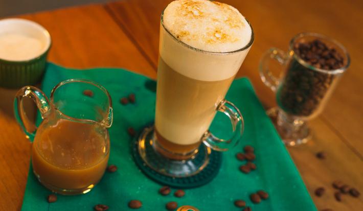 Fotografia em tons de verde de uma mesa de madeira com um paninho verde, uma xícara com a bebida e grãos de café decorando.