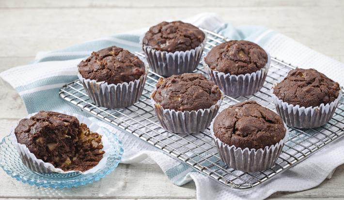 Fotografia em tons de azul em uma mesa de madeira clara com um suporte de ferro com os cupcakes integrais de cacau e banana em forminhas de papel. Ao lado, um pratinho de vidro com um cupcake cortado ao meio.