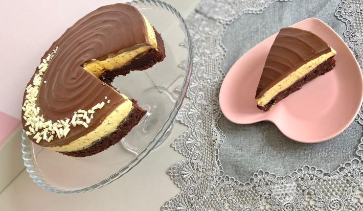 Foto da receita de torta bownie de maracujá, servida em um prato transparente, ao lado de um prato rosa em formato de coração, em uma mesa com uma toalha cinza