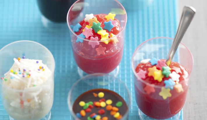 Fotografia em tons de azul em uma bancada cinza com copinhos transparentes contendo a cocadinha, o brigadeiro de colher e o docinho de morango de colher. Ao lado, confeitos coloridos espalhados em formato de estrela.