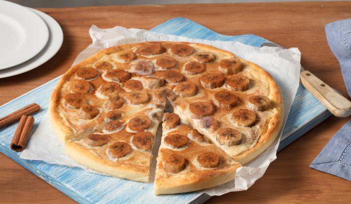 Fotografia em tons de bege e azul, de uma bancada de madeira, ao centro a pizza com rodelas de banana é servida em um papel de seda e uma tábua azul. Ao fundo canela e pratos brancos para decorar.