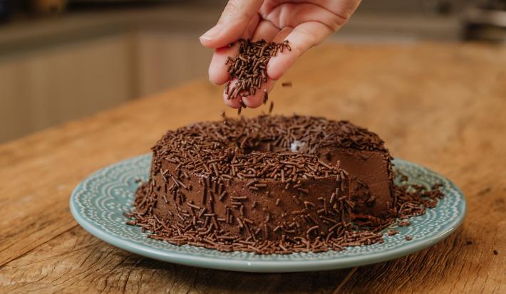 Fotografia em tons de marrom de uma bancada de madeira sobre ela um prato redondo azul com um brigadeirão. Em cima uma mão salpicando granulado de chocolate.