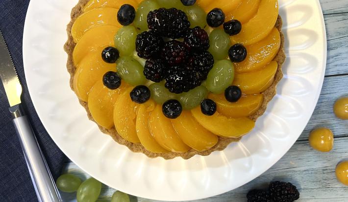 Foto vista de cima da receita de Torta de Frutas com Leite Moça, decorada com pêssegos, uvas, blueberrie, sobre um prato branco em uma bancada de madeira.