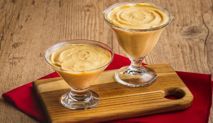 Imagem de duas taças de vidro com a receitas de mousse de maracujá sobre uma tábua de madeira e um pano vermelho.