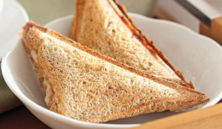 Fotografia em tons de bege em uma mesa com uma toalha bege, um prato redondo fundo branco com dois sanduíches de pão integral tostados e recheados com queijo e lombo.