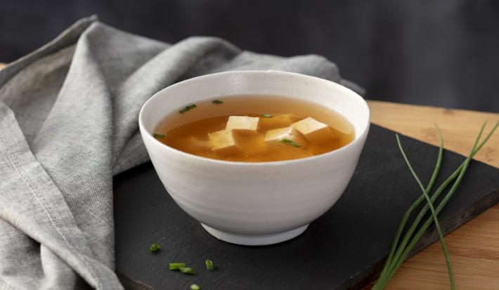 Imagem de uma tigela branca com sopa de missoshiro em cima de uma tábua preta. Ao lado, pedaços de cebolinha.
