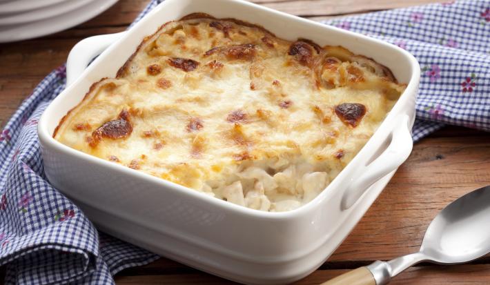 Fotografia em tons de azul em uma bancada de madeira, um pano azul listrado, um recipiente quadrado branco com o frango gratinado dentro dele. Ao lado, uma colher.