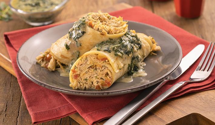 Foto em tons de amarelo da receita de panqueca de frango com molho de espinafre servida em duas porções em um prato cinza sobre um pano vermelho com talheres prateados ao lado. Tudo sobre uma tábua de madeira com uma recipiente ao fundo com o molho
