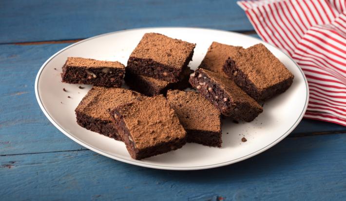 Fotografia em tons de azul, branco e  marrom de uma bancada azul com um prato redondo branco, sobre ele pedaços de brownie de batata-doce. Ao lado um paninho listrado vermelho e branco.