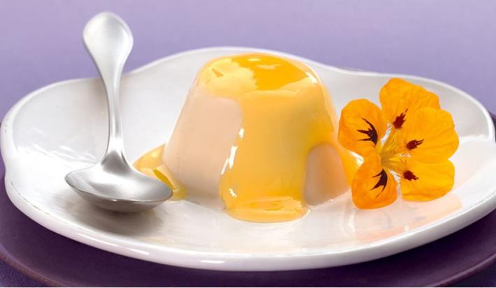 foto em tons lilas e branco. Acima de um prato roxo contém um prato na cor branco que comporta Flan. Ao lado uma colher e uma flor para decorar.o