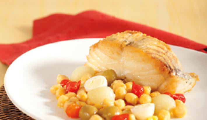 Fotografia em tons de vermelho em uma bancada de madeira clara, um pano vermelho, um jogo americano de vime marrom, um prato redondo raso branco com uma posta de bacalhau com legumes e grão-de-bico ao lado.