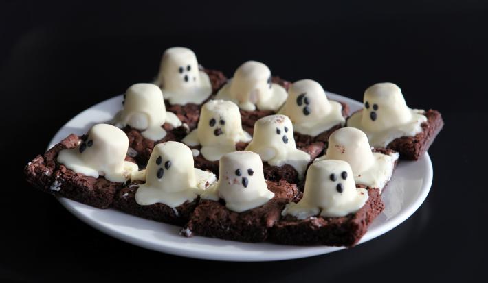 Fotografia de um prato branco com pedaços de brownie de chocolate cortados em quadrados, com marshmallows derretidos em cima representando um fantasma.
