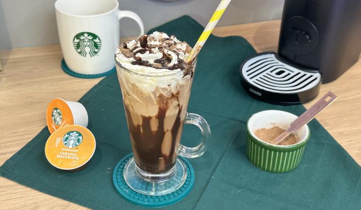 Foto da receita de Frapê de Chocolate. Obseva-se um copo grande decorado com calda de chocolate e chantilly e, ao lado esquerdo, cápsulas de Starbucks.