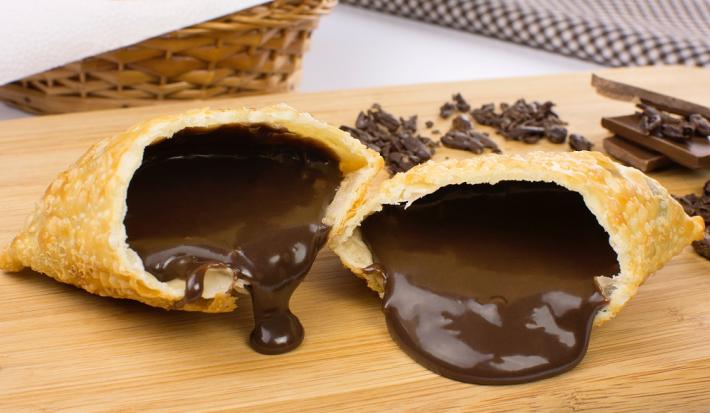 Fotografia em tons de amarelo em uma bancada de madeira de cor branca. Ao centro, há uma tábua de madeira contendo os pasteis e ao fundo há alguns chocolates espalhados e um pano quadriculado preto.